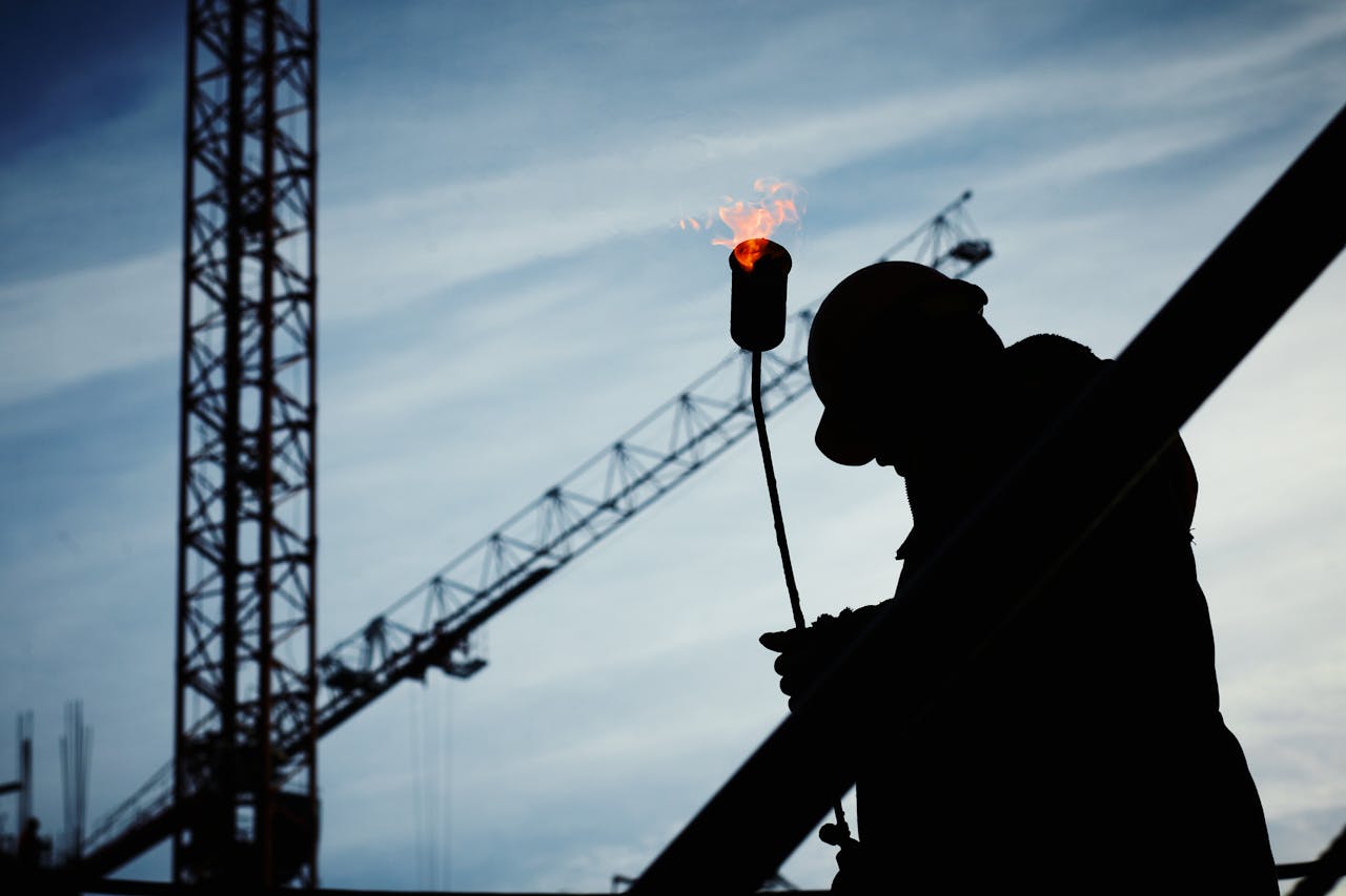 services-img Silhouette of a construction worker using a blowtorch at a building site against a crane-filled skyline.